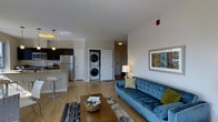 This single-floor residence offers an open-concept layout that seamlessly connects the living area and kitchen, creating a spacious and functional environment. The living area is anchored by a tufted blue sofa paired with a wooden coffee table, complemented by warm wood-look flooring that extends throughout the space. A framed piece of wall art adds a decorative touch, while a table lamp contributes warm ambient lighting.

The kitchen is positioned adjacent to the living area and features dark cabinetry, a stainless steel refrigerator, and a light-toned countertop with bar-height seating, making it ideal for casual dining or entertaining. Pendant lighting above the counter enhances both functionality and style.

A standout feature of this residence is the in-unit stacked washer and dryer, tucked neatly into a dedicated alcove between white panel doors, offering added convenience. Additional doorways visible in the background suggest further rooms beyond the main open area. The white walls and light flooring throughout create a bright, clean aesthetic, while recessed and overhead lighting ensures the space remains well-illuminated.