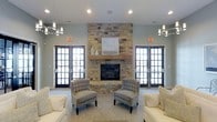 This inviting single-floor space is thoughtfully designed to create a warm and sophisticated atmosphere for its occupants. The centerpiece of the main lounge area is a commanding floor-to-ceiling stone fireplace, crafted from natural stacked stone and topped with a rustic wooden mantel that adds an organic, grounding element to the room. Flanking the fireplace on either side are large, black-framed multi-pane glass doors that flood the interior with generous natural light while offering views to the exterior.

The seating arrangement is both functional and refined, featuring tufted gray accent chairs positioned symmetrically around the fireplace, complemented by cream-toned sofas that create an open and welcoming conversational layout. The neutral color palette of soft grays and warm whites contributes to the space's calm and airy ambiance.

Overhead, contemporary multi-bulb chandeliers provide stylish accent lighting, enhancing the room's modern yet approachable aesthetic. The flooring features a patterned area rug that anchors the seating area, adding texture and visual interest. Smooth, light-gray painted walls complete the clean and polished look of this well-appointed communal gathering space.