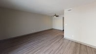 This single-floor residential unit offers a clean, functional layout designed for comfortable everyday living. The spacious living room is a standout feature, showcasing wide-plank luxury vinyl flooring in a cool gray tone, a ceiling fan with light fixture, and a full-width sliding glass door fitted with vertical blinds that flood the room with natural light and provide access to the outdoors. Adjacent to the living area, the galley-style kitchen is efficiently organized with gray cabinetry lining both walls, light-toned countertops, and black appliances including a refrigerator, electric range with overhead exhaust hood, and dishwasher. A rear exterior door provides additional access directly from the kitchen. The unit includes two well-proportioned bedrooms, each featuring the same gray luxury vinyl plank flooring, neutral wall tones, textured ceilings, and individual windows that frame views of lush greenery outside, allowing ample natural light to fill each room. The bathroom offers a generous vanity with an extended countertop, under-sink cabinetry in gray, a large mirror with overhead lighting, and a separate wet area housing a toilet and a full tub-shower combination. Throughout the unit, consistent finishes — including dark baseboards, neutral walls, and textured ceilings — create a cohesive and move-in-ready atmosphere.