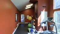 This single-level fitness space is defined by its striking terracotta-red walls, which lend the room a warm, energetic ambiance well-suited to an active environment. The vaulted ceiling adds a sense of openness and height, preventing the space from feeling confined despite its boutique footprint. Overhead fluorescent lighting ensures the area is well-illuminated throughout the day, while windows along the far wall allow natural light to filter in and provide views of the surrounding outdoor area.

The rubber-matted flooring offers a durable, slip-resistant surface appropriate for a variety of workouts. Equipment is thoughtfully arranged throughout the room and includes a treadmill positioned near the entrance, a multi-functional cable or weight machine toward the back, and a dedicated rack holding an assortment of stability balls, medicine balls, and foam rollers. A weight bench with a barbell setup is also present, catering to strength training needs. Informational posters and bulletin boards are mounted on the walls, providing workout guidance. A structural column near the center of the room subtly divides the space into distinct workout zones.