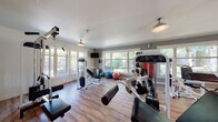 Located at 20699 NE Glisan St in Fairview, Oregon, this single-floor fitness center is a well-appointed facility designed to support a comprehensive workout experience. The space features warm hardwood-style flooring throughout, complemented by neutral gray walls and pendant-style ceiling lights that create an inviting, well-lit atmosphere.

The cardio zone is equipped with multiple treadmills, elliptical machines, and a stationary bike, providing ample options for aerobic training. Adjacent to the cardio area, a strength training section houses a variety of cable machines, weight benches, and multi-use cable systems, including a Paramount cable rack, offering users diverse resistance training options. A dedicated free weights area features a full dumbbell rack for additional versatility.

A standout feature of the space is a functional training and stretching zone stocked with stability balls in multiple sizes, medicine balls, and wall-mounted mats, catering to flexibility, balance, and core conditioning. Large windows along multiple walls flood the interior with natural light and offer pleasant views of the surrounding greenery, enhancing the overall workout environment.

A distinctive corrugated metal partition wall with a sliding barn-style wood door divides the space, adding an industrial-modern design element. A wall-mounted television is also present, providing entertainment during workouts. The facility is thoughtfully organized to maximize usability while maintaining an open, spacious feel.