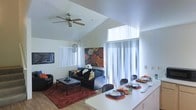 This two-story space showcases an open-concept layout that seamlessly connects the living and dining areas, creating a fluid and functional environment. The living area features vaulted ceilings that lend a sense of spaciousness and volume, enhanced by a ceiling fan that adds both practicality and a casual aesthetic. Natural light pours in through multiple windows fitted with vertical blinds, as well as through elevated clerestory windows positioned high on the wall, flooding the space with brightness throughout the day.

The living area is furnished with dark upholstered seating arranged around a sleek coffee table, anchored by a bold red area rug that adds warmth and color contrast. A vibrant floral artwork on the wall serves as a striking focal point, complementing the contemporary décor. A decorative pendant light adds a touch of character to the space.

Adjacent to the living area, the dining space features a white countertop-style table with seating for multiple guests, set with coordinated place settings in blue and red tones. The open sightlines between both areas make the combined space feel expansive and well-suited for both daily living and entertaining.