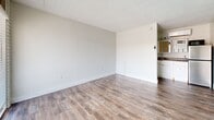 This compact single-floor space is thoughtfully laid out across two areas, offering a functional and clean living environment. The main living area is bright and open, featuring durable wood-look flooring in a warm gray-brown tone that runs throughout. A textured ceiling adds classic character overhead, while crisp white walls and wide baseboards keep the aesthetic clean and neutral. A large sliding glass door paired with a full-height entry door floods the space with natural light and provides views of the surrounding exterior. Roman shades offer light control and a touch of warmth to the entryway. One wall features a distinctive large-format tile panel, adding subtle texture and visual interest. A glimpse into an adjacent area reveals white cabinetry and a sink, suggesting a kitchenette or utility space integrated into the layout. The bathroom is finished with bright white square subway tile wrapping the full tub surround, maintaining the clean, minimal aesthetic found throughout the space. A towel bar and toilet paper holder complete the bathroom's practical fixtures. Overall, the space presents a bright, well-maintained interior with consistent finishes that create a cohesive and functional environment.