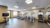 This fitness center, located in Denver, CO, is a single-floor exercise facility designed to support a comprehensive workout experience. The exercise room is thoughtfully organized to maximize the use of its space, offering a wide variety of equipment suited for both cardio and strength training.

The cardio section features multiple treadmills equipped with individual display screens, elliptical cross-trainers, stair climbers, and stationary cycling bikes, providing ample options for aerobic exercise. A rowing machine rounds out the cardio offerings. For strength and functional training, the facility includes medicine balls and additional equipment visible along the walls.

The room is finished with durable rubber sport flooring, ideal for high-activity use, and neutral-toned walls that create a clean, focused environment. A drop ceiling with recessed panel lighting ensures bright, even illumination throughout the space. Large wall-mounted mirrors along one side visually expand the room and assist users in monitoring their form during workouts.

Natural light enters through windows fitted with roller shades, adding to the welcoming atmosphere. A full-length mirrored wall further enhances the sense of openness. Access to the facility is provided through both an interior door and a glass-paneled exterior entry door. Overall, this fitness center offers a practical, well-maintained environment conducive to a productive workout routine.