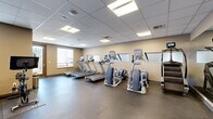 This fitness center, located in Denver, CO, is a single-floor exercise facility designed to support a comprehensive workout experience. The exercise room is thoughtfully organized to maximize the use of its space, offering a wide variety of equipment suited for both cardio and strength training.

The cardio section features multiple treadmills equipped with individual display screens, elliptical cross-trainers, stair climbers, and stationary cycling bikes, providing ample options for aerobic exercise. A rowing machine rounds out the cardio offerings. For strength and functional training, the facility includes medicine balls and additional equipment visible along the walls.

The room is finished with durable rubber sport flooring, ideal for high-activity use, and neutral-toned walls that create a clean, focused environment. A drop ceiling with recessed panel lighting ensures bright, even illumination throughout the space. Large wall-mounted mirrors along one side visually expand the room and assist users in monitoring their form during workouts.

Natural light enters through windows fitted with roller shades, adding to the welcoming atmosphere. A full-length mirrored wall further enhances the sense of openness. Access to the facility is provided through both an interior door and a glass-paneled exterior entry door. Overall, this fitness center offers a practical, well-maintained environment conducive to a productive workout routine.