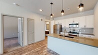 This single-floor, one-bedroom, one-bathroom unit in Colorado Springs, CO offers a well-designed layout that maximizes its compact footprint. The open-concept kitchen and living area serves as the heart of the space, featuring light wood-style flooring that flows throughout. The kitchen is equipped with stainless steel appliances, including a refrigerator, electric range, and microwave, complemented by crisp white cabinetry with black hardware and a tile backsplash. A central island with a warm-toned countertop and an undermount sink doubles as a breakfast bar, illuminated overhead by a trio of stylish pendant lights. The living area receives natural light through windows that overlook the surrounding neighborhood. The bedroom features soft carpeting, a window with a blind, and access to a dedicated closet. The bathroom is finished with wood-style flooring, a dark-toned vanity with a white countertop, and a tiled tub-shower combination. An in-unit laundry area adds everyday convenience. Neutral tones and modern finishes give the entire space a clean, contemporary character.