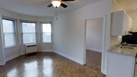This single-floor residence showcases a blend of classic design and practical functionality. The main living area is immediately distinguished by its rich parquet hardwood flooring, which adds warmth and character to the space. A ceiling fan with an integrated light fixture is centrally positioned overhead, providing both air circulation and ambient lighting. Neutral white walls throughout create a clean, versatile backdrop that complements the traditional flooring. Natural light filters in through multiple windows fitted with blinds, illuminating the room and offering views of the surrounding exterior. A baseboard radiator beneath the windows provides in-unit heating. The living area connects seamlessly through a wide doorway to an adjoining room, suggesting an open and flowing layout. Crisp white trim and baseboards frame the space, adding a polished, finished look to the interior. The overall ambiance of the residence is understated and timeless, with classic architectural details that provide a solid foundation for a variety of interior styles.