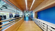 This single-level fitness center combines modern design with functional layout, creating an inviting and well-organized workout environment. The entry corridor sets the tone with warm bamboo flooring and matching wood-paneled walls and ceiling, accented by sleek linear lighting overhead. A glass partition wall along the corridor offers a clear sightline into the main exercise floor, while a built-in storage unit with multiple drawers lines the opposite wall, providing practical organization for equipment and accessories.

The main workout area features an industrial aesthetic with exposed concrete ceilings, structural columns, and visible ductwork, lending the space a loft-like, open feel. Large windows along the exterior walls flood the floor with natural light and offer views of surrounding greenery. Bold blue accent walls punctuate the interior, adding a vibrant, energetic contrast to the neutral tones.

The facility is comprehensively equipped with a full range of cardio machines — including treadmills, ellipticals, and stationary bikes — alongside a diverse selection of strength training equipment such as cable machines, weight racks with dumbbells, and multi-use resistance stations. A dedicated free weights and functional training zone features floor-to-ceiling mirrors, TRX suspension straps, and a workout bench. Multiple wall-mounted TVs are positioned throughout for entertainment during workouts.