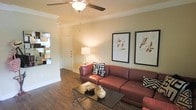This single-story space features a welcoming living area that combines classic architectural details with a warm, inviting ambiance. The room is finished with rich hardwood-style flooring that runs throughout, adding warmth and continuity to the space. Crown molding frames the ceiling, lending an elegant touch to the interior, while a ceiling fan with a built-in light fixture provides both comfort and illumination.

The living area is generously sized, offering ample room for comfortable seating and entertaining. Neutral-toned walls serve as a versatile backdrop, complemented by warm lighting from a table lamp that enhances the cozy atmosphere. Framed artwork adorns the walls, adding a decorative focal point to the space.

A direct entry foyer is visible from the living area, providing a clear and functional transition from the entrance into the main living space. The layout appears open and connected, allowing natural sightlines across the room. Overall, the space presents a well-maintained interior with thoughtful finishing details that contribute to a comfortable and livable environment.