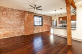 This single-floor urban loft masterfully combines historic industrial character with modern design. Exposed red brick walls stretch throughout the living and bedroom areas, paired with original heavy timber beams and columns that speak to the building's storied past. Rich, dark hardwood flooring runs seamlessly across the open living space, reflecting natural light that pours in through large, architecturally framed windows offering views of the surrounding cityscape.

The open-plan living area is anchored by a ceiling fan and features dramatic exposed ductwork, reinforcing the loft's industrial aesthetic. A generously sized kitchen island with granite countertops serves as the centerpiece of the kitchen, illuminated by a row of pendant lights. Warm wood cabinetry, stainless steel appliances — including a range, microwave, and refrigerator — and tile flooring complete the well-appointed culinary space.

The bedroom areas continue the exposed brick motif, with arched window surrounds adding architectural detail. Closet space includes built-in shelving for added functionality. The bathroom features full tile surrounds in the tub-shower combination, tile flooring, granite countertops, and wood cabinetry, providing a clean and polished finish. This loft offers a compelling balance of raw, historic texture and refined modern living.