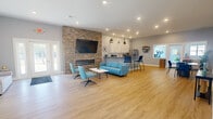 This single-floor community space offers an open, airy layout finished with warm light oak hardwood flooring and soft gray walls illuminated by recessed lighting throughout. The main gathering area is anchored by a striking floor-to-ceiling stacked stone accent wall housing a linear electric fireplace and a mounted flat-screen TV, creating a welcoming focal point. Comfortable seating arrangements, including a blue sofa and coordinating swivel chairs, invite relaxed socializing and casual gatherings. Natural light streams in through multiple windows and a set of glass double entry doors, enhancing the bright and open ambiance. The kitchen area is well-equipped with white shaker-style cabinetry, granite countertops, stainless steel appliances including a side-by-side refrigerator and microwave, and a peninsula with bar seating — ideal for casual dining or entertaining. A dedicated coffee bar station adds a thoughtful amenity. The entrance flows seamlessly into the main lounge, where additional seating areas provide flexible use of the space. The accessible bathroom is outfitted with stainless steel grab bars and ADA-compliant fixtures, ensuring inclusive usability. A window opening to a fitness area with exercise equipment rounds out the functional offerings of this thoughtfully designed space.