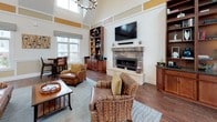 Located at 191 Beach in Salisbury, Massachusetts, this single-floor community space offers a thoughtfully designed interior that blends comfort and functionality with coastal charm. The centerpiece of the space is a grand open living area boasting soaring vaulted ceilings, large windows that invite abundant natural light, and a striking stone fireplace topped by a wall-mounted television. Built-in shelving units flank the fireplace, providing both display and storage, while rich wood-tone flooring runs throughout, anchoring the warm, inviting atmosphere. Comfortable wicker and leather seating arrangements are complemented by a soft area rug, creating an ideal gathering setting. Adjacent to the main living area, a dining nook features a raised round wooden table with cushioned counter-height chairs beneath bright windows, offering views of the surrounding neighborhood. A decorative coastal-themed wreath adorns the entryway door, reinforcing the beachside character of the space. The compact kitchenette is well-appointed with rich cabinetry, dark granite countertops, stainless steel appliances, and ample storage. A secondary seating area near the glass entry doors provides additional flexible space with views to the outdoors. The overall ambiance is warm, coastal, and community-focused, making this a welcoming destination for residents and guests alike.
