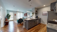 Located at 555 5th Ave. in Columbus, Ohio, this single-floor residence offers a well-designed open-concept layout that seamlessly connects the kitchen, dining, and living areas. The kitchen is a functional centerpiece, featuring gray shaker-style cabinetry, granite countertops, a large island with an undermount sink and pendant lighting, and a full suite of stainless steel appliances. Warm hardwood-style flooring runs throughout the main living areas, adding continuity and a welcoming feel. The living room is bright and airy, benefiting from large windows dressed with sheer curtains that allow generous natural light to filter in. A glass door provides direct access to an outdoor area. The space includes two bedrooms, each carpeted and outfitted with windows that bring in natural light and views of the surrounding greenery. Both bathrooms are finished with large-format tile flooring, dark wood vanities with granite countertops, and glass-enclosed shower/tub combinations. The overall aesthetic throughout the residence is modern and cohesive, with soft gray walls, recessed lighting, and thoughtful design details that create a comfortable, contemporary living environment.