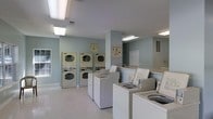 This single-floor shared laundry facility is designed with functionality and ease of use in mind. The space features a generous arrangement of top-loading washing machines lined up in a row, complemented by several stacked washer-dryer units along the walls, providing ample capacity for multiple users at once. The room is finished with light-toned flooring that enhances the sense of cleanliness and openness throughout. Soft, pale blue-green walls contribute to a calm and pleasant atmosphere, making the space feel welcoming rather than purely utilitarian. Overhead fluorescent lighting ensures the entire room is brightly and evenly illuminated, supporting a practical environment at any hour. Informational signage is posted on the walls, offering guidance on machine operation and facility rules. A window along one wall allows for a touch of natural light. The layout is thoughtfully organized, leaving clear pathways between equipment for comfortable movement. The textured ceiling and clean white trim add subtle architectural detail, while the overall design prioritizes efficiency, accessibility, and a tidy, well-maintained environment for all users.