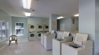This single-floor shared laundry facility is designed with functionality and ease of use in mind. The space features a generous arrangement of top-loading washing machines lined up in a row, complemented by several stacked washer-dryer units along the walls, providing ample capacity for multiple users at once. The room is finished with light-toned flooring that enhances the sense of cleanliness and openness throughout. Soft, pale blue-green walls contribute to a calm and pleasant atmosphere, making the space feel welcoming rather than purely utilitarian. Overhead fluorescent lighting ensures the entire room is brightly and evenly illuminated, supporting a practical environment at any hour. Informational signage is posted on the walls, offering guidance on machine operation and facility rules. A window along one wall allows for a touch of natural light. The layout is thoughtfully organized, leaving clear pathways between equipment for comfortable movement. The textured ceiling and clean white trim add subtle architectural detail, while the overall design prioritizes efficiency, accessibility, and a tidy, well-maintained environment for all users.