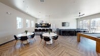 The E-Lounge and Community Area at 1200 Allison Dr in Vacaville, California is a thoughtfully designed, open-concept communal space built for both relaxation and social engagement. The layout flows seamlessly across herringbone-patterned hardwood flooring, connecting distinct functional zones within one expansive, airy interior.

The fully equipped kitchen serves as a central hub, featuring a striking island topped with veined marble-style countertops, a professional-grade gas range, and a premium refrigerator with gold hardware accents. A bold black square-tile backsplash and floating wood shelves add a sophisticated, contemporary edge, while pendant lighting overhead enhances the space's modern aesthetic.

Adjacent to the kitchen, a spacious lounge area offers generous sectional seating arranged around a wall-mounted flat-screen TV, ideal for communal viewing and casual gatherings. Round white tables paired with modern black chairs provide ample dining and social seating throughout the open floor plan. A pool table positioned near floor-to-ceiling glass doors and windows invites recreation while drawing in abundant natural light and outdoor views.

Soaring ceilings with clerestory windows amplify the sense of openness, and a ceiling fan ensures year-round comfort. An interior overlook with mesh railing connects to an adjoining stairwell area, adding vertical dimension and architectural interest to the overall design. The clean white walls and warm flooring create a welcoming, contemporary ambiance throughout.