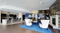 This thoughtfully designed single-floor space combines contemporary interior style with exceptional outdoor access, creating an inviting environment for both relaxation and social gatherings. The interior lounge area is anchored by rich hardwood flooring and a vibrant blue area rug that sets the tone for the space's nautical-inspired palette. A striking woven pendant light serves as a central design focal point, complementing the recessed lighting throughout. Comfortable seating arrangements — including patterned accent chairs and upholstered sofas — are organized around a sleek coffee table, encouraging conversation and leisure. A dark, built-in shelving unit along one wall displays curated décor accents, adding depth and character to the room. Adjacent to the lounge, a fully equipped kitchen and dining area features bar-height seating, dark cabinetry, and modern appliances, making it well-suited for hosting and events. A bold blue accent wall bridges the two zones with a cohesive design thread. Outside, the expansive waterfront terrace offers sweeping river views beneath open skies. Modern wood-and-metal picnic tables and benches provide ample seating, while an outdoor grill station enhances the space's entertaining capabilities. The terrace's open, airy design invites residents and guests to enjoy the scenic waterfront setting year-round.