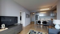 This single-floor unit showcases a thoughtfully designed open-concept layout that seamlessly integrates the living and kitchen areas. The space features light hardwood flooring that runs throughout, contributing to a bright and cohesive aesthetic. Walls are finished in a soft, neutral tone that enhances the overall sense of openness.

The kitchen is outfitted with sleek, light grey cabinetry that extends to the ceiling, providing ample storage. A stainless steel refrigerator anchors the space, complemented by a clean white countertop and a compact kitchen bar with black metal stools — ideal for casual dining. Under-cabinet and pendant lighting ensures the kitchen remains well-lit and functional.

The living area accommodates a media console with a large flat-screen television and is anchored by a patterned area rug, adding a layer of visual interest. Framed artwork adorns the walls, adding a personalized touch to the space.

A hallway leads to the bedroom and a walk-in closet, which is visible through an open door, offering organized hanging storage. A secondary doorway along the hall provides access to an additional room. The overall ambiance is clean, modern, and functional.