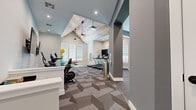 Located at 3788 Old Port Royal Rd N in Spring Hill, Tennessee, this single-story fitness facility offers a thoughtfully designed workout environment across a generously sized open floor plan. The space is defined architecturally by soaring vaulted ceilings accented with rustic wood paneling, pendant lighting, and recessed fixtures that collectively create a bright, energizing atmosphere. Geometric-patterned carpet tiles cover the main cardio and cycling areas, adding visual interest while providing functional underfoot comfort.

The primary workout area houses a row of TRUE-brand treadmills alongside stationary and cycling bikes, all positioned beneath the dramatic cathedral ceiling and adjacent to windows that invite natural light. Wall-mounted flat-screen televisions provide entertainment throughout. A built-in counter station with seating offers a convenient space for tracking workouts or brief rest.

A dedicated strength and functional training zone, finished with rubber flooring for durability, features a heavy bag, battle ropes, a cable machine, a dumbbell rack with a full weight selection, and a weight bench. Wall mirrors enhance this area's utility. An additional mirrored room is visible off the main floor, suitable for stretching or supplemental training. Overall, the facility balances modern design with comprehensive, practical fitness amenities.