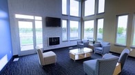 This single-floor lounge space makes an immediate impression with its dramatic two-story wall of windows, flooding the interior with an abundance of natural light and offering open views of the surrounding outdoor landscape. The soaring ceiling height amplifies the sense of spaciousness and grandeur throughout the room.

At the heart of the lounge is a sleek, tile-clad fireplace framed by a light stone surround, providing a refined focal point and a warm atmosphere. A large wall-mounted flat-screen television is positioned above the fireplace, making the space equally suited for casual viewing or presentations.

The seating arrangement is thoughtfully designed for comfort and conversation, featuring a mix of plush armchairs, a sectional-style seat, and a modern wood-and-metal coffee table with open shelving. The upholstered furniture in cool, neutral tones complements the room's contemporary aesthetic. Underfoot, a richly patterned dark carpet adds texture and depth to the space.

Natural light streams in from all angles through the expansive multi-pane windows that line multiple walls, seamlessly connecting the interior to the outdoors. A glass door provides direct access to an exterior area, further enhancing the open, airy feel. The overall ambiance is welcoming, polished, and well-suited for a variety of communal uses.