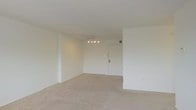 This single-floor residential space offers a clean, open layout designed for comfortable everyday living. The main living area is generously proportioned, featuring wall-to-wall neutral carpet flooring that adds warmth and a sense of continuity throughout the space. Crisp white walls and a white ceiling work together to create a bright, airy atmosphere that feels open and inviting.

A ceiling fan with a multi-bulb light fixture is centrally positioned in the living area, providing both overhead illumination and air circulation. The entryway is cleanly integrated into the living space, with a solid white door that contributes to the room's cohesive, minimalist aesthetic.

The layout makes efficient use of the available footprint, with defined zones that offer flexibility for various furniture arrangements. Subtle architectural details, including column-like wall projections, add a degree of structural character to the otherwise streamlined interior. The overall ambiance is calm and neutral, presenting a versatile blank canvas well-suited for a variety of interior styles and personal touches.