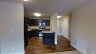 This single-floor space offers a thoughtfully designed layout that balances functionality and modern style. The kitchen serves as the centerpiece of the home, showcasing rich, dark espresso-toned cabinetry that reaches toward the ceiling, providing ample storage. Light granite countertops offer a striking contrast against the dark cabinets, while stainless steel appliances — including a refrigerator, range, and built-in microwave — add a sleek, contemporary finish. A curved kitchen island extends the prep and dining space, making it an ideal spot for casual meals or entertaining. The kitchen flows seamlessly into the adjoining living area, unified by warm hardwood-style flooring that runs throughout. A textured ceiling adds subtle visual interest overhead, while flush-mount lighting ensures the space is well-lit throughout. Neutral, light-toned walls keep the atmosphere open and airy. A hallway visible from the kitchen leads to additional rooms, with clean white doors and crisp trim detailing that maintain a polished, cohesive aesthetic throughout the home. The transition from hard flooring to soft carpet hints at a comfortable living or bedroom area beyond, completing a well-rounded and inviting residential space.