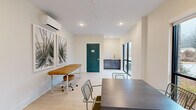 This single-room professional space is thoughtfully designed with a clean, modern aesthetic that balances functionality and comfort. The light-toned hardwood flooring runs throughout, lending warmth to the otherwise crisp, neutral interior. Recessed lighting is complemented by abundant natural light streaming in through multiple large, black-framed floor-to-ceiling windows, offering views of the surrounding neighborhood and greenery outside.

A wall-mounted mini-split system provides efficient climate control, positioned above a pair of framed botanical art prints that add a natural, calming touch to the space. A treatment or work table with a rolling stool occupies one side of the room, while a central table with wire-frame chairs suggests flexible use for consultations, meetings, or collaborative work.

Toward the rear of the room, a teal two-panel accent door provides a bold pop of color against the warm white walls, leading to an adjoining space. Adjacent to this door is a built-in dark-cabinetry wet bar or utility sink area, offering practical functionality. A sliding glass door on the opposite wall provides additional natural light and outdoor access.

The overall ambiance is bright, clean, and professional — well-suited for a wellness studio, therapy office, or boutique professional practice.