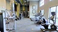 This modern fitness center is designed to accommodate a comprehensive workout experience within a single, open-plan floor. The space features soaring ceilings that lend an airy, expansive feel, enhanced by large windows that invite abundant natural light throughout the room. A row of up-to-date cardio machines, including treadmills and ellipticals equipped with individual digital displays, lines one side of the facility, offering members a well-organized and efficient workout zone. On the opposite side, a dedicated strength training area houses a variety of cable and resistance machines suited for full-body conditioning. Full-length mirrors along the walls amplify the sense of space and allow exercisers to monitor their form. The flooring features patterned carpet tiles that add a contemporary touch while providing comfort underfoot. A striking large ceiling fan serves as both a functional and architectural focal point overhead. Framed art pieces adorn the neutral-toned walls, contributing a polished, welcoming ambiance. Accents of bold green and dark paneling add visual interest and a modern edge to the overall design, making this fitness center as aesthetically appealing as it is functional.