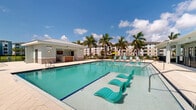 Located at Savona Point in Cape Coral, Florida, this single-story community amenity facility offers a well-appointed outdoor living experience designed for relaxation and socializing. At the heart of the space is a resort-style swimming pool surrounded by a spacious tiled pool deck, complete with lounge chairs and colorful umbrellas, all enclosed by a sleek black iron fence. Towering palm trees and manicured green lawns frame the area, providing a lush, tropical backdrop under the Florida sun. A generously sized covered lanai extends along the pool's edge, outfitted with ceiling fans to ensure comfort in the warm climate. The lanai features multiple seating arrangements, including wicker lounge chairs and conversation sets with coordinating cushions, ideal for casual gatherings. An outdoor kitchen area is equipped with stone countertops, stainless steel cabinetry, bar seating, a wall-mounted television, and a built-in grill, making it a fully functional entertaining space. Accessible restroom facilities are conveniently located on the pool deck. Scenic views of the adjacent waterway and the surrounding residential buildings complete the setting, creating a cohesive and inviting community retreat.