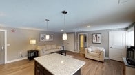 This single-floor space is designed around an open-concept layout that seamlessly connects the kitchen and living areas, creating a sense of spaciousness and ease of flow. The kitchen is anchored by a substantial island topped with light granite countertops, complemented by dark cabinetry beneath. Two pendant lights with brushed nickel fixtures hang above the island, providing focused task lighting while adding a modern touch.

The living area extends naturally from the kitchen, featuring warm-toned laminate hardwood flooring that runs throughout the space. Neutral gray walls provide a clean, contemporary backdrop for the cream-colored seating arrangements, which include a full sofa and an accent chair. A textured area rug anchors the seating area, while decorative wall art adds visual interest.

Recessed lighting supplements the pendant fixtures overhead, and a textured ceiling adds subtle dimension. A doorway visible in the background leads to additional rooms, suggesting a functional layout beyond the main living space. Overall, the space presents a cohesive, neutral palette with modern finishes that create a comfortable and inviting atmosphere.