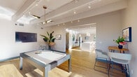 This thoughtfully designed single-floor space combines modern aesthetics with functional versatility across several distinct yet interconnected areas. Exposed structural beams run the length of the vaulted ceilings throughout, lending an open, loft-like character to the interior, while warm light wood flooring unifies the layout seamlessly.

The game room serves as a welcoming entry point, anchored by a sleek contemporary pool table beneath a sculptural multi-arm chandelier. A wall-mounted flat-screen display, curated artwork, and a tall indoor plant add personality to the bright white walls, which are further animated by track lighting overhead.

The space flows naturally into a striking open kitchen and gathering area, defined by a row of dramatic arched windows that frame lush outdoor views and flood the interior with natural light. A long kitchen island with a textured stone-finished base and a crisp white countertop serves as the centerpiece, accompanied by modern pendant lighting and a set of stylish bar seating. A secondary prep and service counter with a round mirror and dark sink adds both function and visual interest.

Adjacent to the kitchen, the lounge area offers a comfortable retreat with a bold orange sectional sofa, organic-shaped wooden coffee tables, and a large wall-mounted screen — all bathed in sunlight streaming through the arched windows. The overall ambiance is bright, contemporary, and inviting.