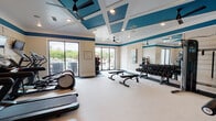 This fitness center is a thoughtfully designed workout facility that combines functionality with polished aesthetic appeal. The space is defined by its striking coffered ceiling, painted in crisp white and accented with bold teal panels, which adds a sense of architectural depth and visual interest overhead. Ceiling fans are integrated throughout, ensuring comfortable airflow during workouts.

The facility is divided into well-organized zones to accommodate a variety of training styles. A dedicated cardio area lines one wall, featuring multiple modern treadmills and an elliptical machine, each equipped with individual display screens. Wall-mounted flat-screen televisions are positioned directly in front of the cardio equipment for entertainment during workouts. Adjacent to this section is a free weights area, complete with a full rack of dumbbells and adjustable workout benches, backed by a large floor-to-ceiling mirror that amplifies the sense of space. A cable machine and additional strength training equipment occupy a separate zone, offering a comprehensive range of resistance training options.

Large sliding glass doors and wide windows flood the interior with natural light and provide open views of the outdoor surroundings. The neutral, light-toned flooring transitions to darker rubber matting in high-use zones, balancing practicality with design. Decorative artwork adorns the walls, lending a refined touch to the overall atmosphere.