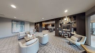This elegantly designed single-floor amenity lounge offers a seamlessly integrated layout organized into several functional zones. The main lounge area features plush, light-toned curved and angular seating arrangements centered around mirrored coffee tables, all anchored by a bold geometric-patterned carpet that runs throughout the space. Recessed lighting illuminates the room evenly, complementing the neutral grey and deep espresso-brown color palette.

A striking dark wood open-shelving partition divides the main lounge from the business center, where multiple computer workstations are set against a dramatic backlit, textured metallic accent wall adorned with cascading pendant lighting. Adjacent to the business center, a dining-height table with upholstered chairs provides a flexible conference or collaborative workspace.

A dedicated media room offers wall-mounted television viewing with streamlined grey armchairs and direct access through glass doors to an exterior terrace area. Nearby, a kitchenette alcove features dark cabinetry, a stainless steel under-counter refrigerator, and a textured tile backsplash, providing convenient refreshment capabilities. The overall ambiance is polished and contemporary, suited for both focused work and relaxed socializing.