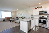 This single-floor residence offers a thoughtfully designed layout that balances functionality with modern style. The open-concept kitchen is a standout feature, showcasing crisp white shaker cabinetry, granite-patterned countertops, stainless steel appliances including a range, microwave, and refrigerator, and rich dark hardwood-style flooring. A peninsula with pendant lighting and bar seating provides a natural gathering point that connects the kitchen to the main living area. The living room is warm and inviting, finished with plush carpeting and large windows that allow abundant natural light to filter in, offering views of the surrounding greenery. Recessed lighting throughout enhances the bright, airy atmosphere. The bedroom is generously proportioned, featuring soft carpet, a ceiling fan, and a window with outdoor views, providing a comfortable and restful retreat. A spacious walk-in closet with dual hanging rods and shelving on multiple walls offers excellent storage capacity. The bathroom is well-appointed with a white vanity, granite-style countertop, wood-look flooring, and a full tub-shower combination. Clean neutral tones and white trim carry consistently throughout the space, lending a cohesive and polished aesthetic overall.