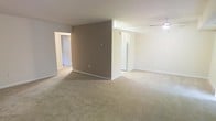 This single-floor residence features an open-concept main living area finished with wall-to-wall carpet in a warm beige tone, creating a comfortable and cohesive feel throughout the space. A prominent taupe accent wall serves as a central architectural element, visually dividing the layout while maintaining an open flow between areas. The neutral white walls and trim throughout contribute to a clean, bright aesthetic.

A ceiling fan with a light fixture is installed in one section of the living area, providing both air circulation and ambient lighting. Natural light enters through a window or glass door visible beyond the accent wall, offering a glimpse of an outdoor area and adding brightness to the interior.

Hallways extend from the main living space, leading to additional rooms, suggesting a functional floor plan with dedicated private areas. White baseboards line the perimeter of each room, providing a crisp contrast to the carpeted floors. The overall ambiance is understated and move-in ready, with a straightforward layout suited to a variety of furnishing arrangements.