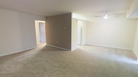 This single-floor residence features an open-concept main living area finished with wall-to-wall carpet in a warm beige tone, creating a comfortable and cohesive feel throughout the space. A prominent taupe accent wall serves as a central architectural element, visually dividing the layout while maintaining an open flow between areas. The neutral white walls and trim throughout contribute to a clean, bright aesthetic.

A ceiling fan with a light fixture is installed in one section of the living area, providing both air circulation and ambient lighting. Natural light enters through a window or glass door visible beyond the accent wall, offering a glimpse of an outdoor area and adding brightness to the interior.

Hallways extend from the main living space, leading to additional rooms, suggesting a functional floor plan with dedicated private areas. White baseboards line the perimeter of each room, providing a crisp contrast to the carpeted floors. The overall ambiance is understated and move-in ready, with a straightforward layout suited to a variety of furnishing arrangements.