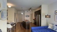 This single-floor space is designed with an open-concept layout that seamlessly connects the living and kitchen areas, creating a cohesive and functional environment. The kitchen is outfitted with stainless steel appliances, including a full-size refrigerator, microwave, and range, all complemented by dark cabinetry that extends to the upper walls for generous storage. A central kitchen island with a dark finish serves as both a prep space and casual dining area, accompanied by a bar-height stool. Pendant lighting hangs above the island, adding focused illumination, while a track lighting system overhead provides additional flexibility. The rich, dark hardwood flooring runs throughout the space, grounding the design and adding warmth. The living area, partially visible, features a bold blue sofa that introduces a vibrant accent against the neutral white walls. Decorative oval wall art adds a subtle artistic touch to the dining-adjacent wall. Doors leading to additional rooms are visible along the perimeter, suggesting a well-organized floor plan. The overall ambiance is modern and urban, with a clean aesthetic balanced by warm tones.