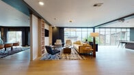 Located in the vibrant South End neighborhood of Charlotte, NC, this sophisticated single-floor amenity space blends contemporary design with refined functionality. Light-toned hardwood flooring flows seamlessly throughout, unifying the various zones and lending warmth to the modern aesthetic.

The main lounge area is anchored by a striking circular pendant light and a built-in shelving wall in natural wood, paired with a wall-mounted display. Expansive floor-to-ceiling windows flood the space with natural light and offer open views of the surrounding Charlotte skyline and greenery, creating an airy, inviting atmosphere.

The dining area features a large round table with upholstered chairs beneath a bold, oversized drum pendant, complemented by framed artwork on the walls. A separate dining zone with sliding glass doors and dramatic draping curtains provides additional seating capacity, ideal for gatherings or events.

A kitchenette is outfitted with dark wood cabinetry, a white quartz countertop, stainless steel appliances, and a stunning bold geometric black-and-white tile feature wall that serves as a visual centerpiece. An adjoining conference or private meeting room rounds out the layout, offering a more intimate setting. Throughout, the space balances bold design accents with clean, open interiors.