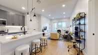 This well-appointed single-floor residence showcases a thoughtfully designed open-concept layout that seamlessly connects the kitchen, living, and dining areas. The kitchen is equipped with gray shaker-style cabinetry, stainless steel appliances including a french-door refrigerator and gas range, white quartz countertops, and a peninsula with bar seating beneath striking matte black pendant lights. Light hardwood flooring flows throughout the main living area, which offers generous room for both lounging and a dedicated workspace, anchored by a wall-mounted TV and access to a glass-door balcony that invites natural light and city views.

Both bedrooms are carpeted and feature large windows with urban skyline views, ceiling fans or accent lighting, and access to closet storage. The primary bathroom is finished with large-format light tile flooring, gray cabinetry, a white quartz vanity, matte black fixtures, and an illuminated mirror, with a walk-in closet accessible from within. A dedicated laundry area houses a full-size washer and dryer with built-in shelving above. Throughout, a neutral palette of whites and grays creates a clean, contemporary ambiance.