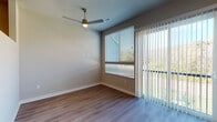 This single-floor residence offers a clean, contemporary design highlighted by consistent wood-style flooring throughout the main living areas. The open living space is filled with natural light streaming through large windows fitted with horizontal blinds and a sliding glass door framed by vertical blinds, providing direct outdoor views of surrounding greenery. A modern ceiling fan with an integrated light fixture overhead enhances airflow and adds a sleek, functional detail to the room.

The kitchen is efficiently laid out along a single wall, featuring light wood cabinetry with brushed metal hardware, granite-style countertops, and a tiled backsplash. A full suite of stainless steel appliances—including a refrigerator with ice and water dispenser, electric range, microwave, and dishwasher—provides practical functionality. An adjacent area with elevated ceilings connects the kitchen to the living space, adding a sense of openness.

The bathroom includes a granite-top vanity with an under-mount sink, light wood cabinetry, a large mirror, and a full tub-shower combination with tiled surrounds and built-in shelving. A separate carpeted room offers a quieter, softer space suitable for use as a bedroom, complete with neutral tones and ample wall space.