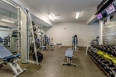 Located at 8610 N Kansas Ave in Kansas City, Missouri, this fitness center offers a comprehensive workout facility designed to accommodate a wide range of exercise needs. The space is organized across a single floor and divided into distinct functional zones, each tailored to a specific type of training.

The cardio zone features multiple treadmills and elliptical machines, including Precor equipment, positioned along a wall of large glass doors that open to an outdoor view, allowing natural light to fill the area and create an energizing atmosphere. Mounted flat-screen televisions above the cardio equipment provide entertainment during workouts.

Adjacent to the cardio area, a dedicated strength and free weights section houses a full dumbbell rack with yellow-accented storage, multiple weight benches, and Nautilus cable and plate-loaded machines. Full-length mirrors line the walls, enhancing the sense of space and allowing users to monitor their form.

A separate strength training room provides additional resistance machines and functional training equipment, finished with durable rubber flooring throughout. The facility also includes a stretching and utility area with accessible storage and sanitation stations.

The overall design features neutral gray walls, recessed and fluorescent lighting, and carpet-tiled flooring in the main corridors, creating a clean, modern, and functional environment suited for serious fitness training.