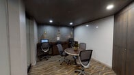 Located at 3700 Cole Ave in Dallas, Texas, this single-floor business center is thoughtfully designed to support both focused work and collaborative gatherings. The main workspace features a built-in wooden desk along one wall, equipped with two computer monitors and a shared printer, all illuminated by a trio of decorative gold pendant lights that hang against a textured slate-toned accent wall. Ergonomic mesh chairs provide comfortable seating at each workstation. A wave-patterned carpet in warm earth tones anchors the room and adds a layer of visual sophistication. Opposite the workstations, a round wooden table with a small floral centerpiece is surrounded by additional mesh chairs, creating an inviting spot for informal meetings or group collaboration. A smaller adjoining area offers a more intimate setting, highlighted by a rich dark wood built-in cabinet and an eye-catching wall display of decorative sculptural plates in navy and gold. The hallway connecting the spaces features wood-look tile flooring in soft taupe tones and recessed lighting, contributing to a clean and professional aesthetic. Recessed ceiling lights throughout the space ensure even, functional illumination. The overall ambiance is contemporary and polished, well-suited for productivity in a community-oriented setting.