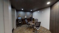 Located at 3700 Cole Ave in Dallas, Texas, this single-floor business center is thoughtfully designed to support both focused work and collaborative gatherings. The main workspace features a built-in wooden desk along one wall, equipped with two computer monitors and a shared printer, all illuminated by a trio of decorative gold pendant lights that hang against a textured slate-toned accent wall. Ergonomic mesh chairs provide comfortable seating at each workstation. A wave-patterned carpet in warm earth tones anchors the room and adds a layer of visual sophistication. Opposite the workstations, a round wooden table with a small floral centerpiece is surrounded by additional mesh chairs, creating an inviting spot for informal meetings or group collaboration. A smaller adjoining area offers a more intimate setting, highlighted by a rich dark wood built-in cabinet and an eye-catching wall display of decorative sculptural plates in navy and gold. The hallway connecting the spaces features wood-look tile flooring in soft taupe tones and recessed lighting, contributing to a clean and professional aesthetic. Recessed ceiling lights throughout the space ensure even, functional illumination. The overall ambiance is contemporary and polished, well-suited for productivity in a community-oriented setting.