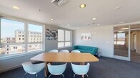Located at 3009 M E Valentine Dr in Raleigh, NC, this single-floor space is designed to support both collaborative work and relaxed gathering. The layout is divided into two primary zones: a conference area and a lounge seating area, unified by consistent modern design elements throughout.

The conference area is anchored by a sleek, wood-toned oval table surrounded by contemporary white chairs. Large windows wrap around multiple walls, flooding the space with natural light and offering open views of the surrounding Raleigh cityscape. Recessed ceiling lighting complements the natural light, ensuring a well-lit environment at all hours.

The lounge area features a bold teal tufted sofa and matching teal accent chairs paired with a minimalist round side table, providing a welcoming space for informal meetings or relaxation. A dark charcoal accent wall adds depth and contrast to the otherwise light gray and white walls. Colorful wall art, including botanical prints, adds visual interest throughout the space.

The flooring is covered in deep navy-blue carpet, grounding the vibrant furnishings and contributing to the space's cohesive, professional aesthetic. Glass-paneled interior doors connect the zones while maintaining an open, airy feel. Overall, the space strikes a balance between functional meeting infrastructure and comfortable, design-forward interiors.