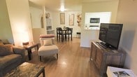 This single-story space is thoughtfully designed with an open-concept floor plan that encourages fluid movement between the living, dining, and kitchen areas. Warm hardwood flooring runs throughout, lending a cohesive and inviting character to the entire space. The living area is comfortably arranged with accent seating, including a cushioned slipper chair and a rustic wooden media console that anchors the entertainment setup. Decorative touches such as framed wall art and woven basket accents add a personalized, homey feel. The dining area features a dark wood high-top table paired with matching bar-height chairs, creating a casual yet defined space for meals. A pass-through window connects the dining area to the kitchen, which is equipped with white cabinetry, a built-in microwave, and ample counter space, contributing to an efficient and bright cooking environment. A secondary room visible in the background mirrors the white cabinetry aesthetic, suggesting additional storage or utility space. Overall, the layout is practical and well-organized, with a warm, neutral color palette that creates a comfortable and welcoming ambiance throughout.
