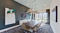 This professionally designed conference room offers a polished and contemporary environment ideal for meetings and collaborative work. The space is anchored by a generously sized dark wood boardroom table surrounded by ergonomic tan leather chairs with chrome bases, comfortably accommodating a full team. Soaring ceilings lend an impressive sense of volume and openness to the room, enhanced by a pair of striking brushed metallic linear pendant fixtures that cast a warm, focused light over the work surface. One wall is finished in a deep charcoal gray, serving as a dramatic backdrop for a wall-mounted flat-screen display and a white credenza — a refined combination that balances function with style. Curated abstract artwork adorns the walls, adding a creative and sophisticated character to the professional setting. Floor-to-ceiling windows along one side of the room flood the interior with natural light and frame views of an outdoor courtyard with landscaping and architectural elements beyond. Sheer drapery panels provide adjustable light control while maintaining the bright, airy atmosphere. The floor is covered in a richly patterned geometric carpet in deep navy and gray tones, grounding the space with texture and visual interest. A framed monochromatic photographic print completes the art collection, contributing to the room's modern, gallery-like ambiance.