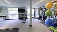 This fitness center is a thoughtfully designed, single-floor space built to support a comprehensive workout experience. The layout is open and spacious, allowing for easy movement between stations and equipment zones. Large windows line the perimeter walls, flooding the interior with abundant natural light and offering views of the surrounding outdoors, creating an energizing atmosphere for exercise.

The strength training area is well-appointed, featuring a multi-tiered dumbbell rack stocked with a full range of free weights, an adjustable workout bench, and a cable machine system suitable for a wide variety of resistance exercises. A wall-mounted flat-screen television is positioned above the free weight station, providing entertainment during workouts. Flooring transitions between standard and rubberized surfaces in the lifting zones, offering durability and safety where needed.

A structural column is integrated into the floor plan, subtly dividing the space without disrupting its open feel. Recessed and linear LED lighting supplements the natural light from the windows, ensuring the space remains consistently well-lit. The neutral color palette—light walls, light flooring, and dark equipment—contributes to a clean, professional aesthetic. Overall, this fitness center delivers a functional and inviting environment designed to accommodate a range of fitness activities.
