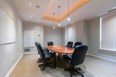 Located at 65 Times Ave in Markham, Ontario, this professional conference room offers a well-appointed setting designed for focused meetings and collaborative work. The room's most distinctive architectural feature is its recessed tray ceiling, finished with warm amber accent lighting that adds a polished, inviting ambiance to the otherwise neutral-toned space. Complementing the tray ceiling are recessed pot lights and sleek pendant lights that hang above the central oval conference table, ensuring the workspace is well-illuminated throughout.

The oval table, finished in warm wood tones, is surrounded by six black leather executive chairs, providing comfortable seating for small team meetings or client discussions. Neutral grey walls and light-toned carpeting contribute to the room's clean, professional atmosphere. Multiple windows fitted with vertical blinds line the walls, allowing for adjustable natural light while maintaining privacy.

Additional functional elements include a wall-mounted whiteboard for presentations and brainstorming sessions, built-in storage cabinetry and a hutch unit along one wall offering space for office equipment and supplies, and a dedicated entry door. The room's compact yet efficient layout makes it a practical and comfortable space for professional gatherings at this Markham address.