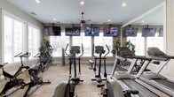 This single-floor fitness center offers a comprehensive workout facility designed to accommodate a wide range of exercise needs. The space is thoughtfully organized into distinct zones, beginning with a cardio area outfitted with treadmills, elliptical trainers, and stationary bikes arranged along large windows that flood the room with natural light and provide views of the surrounding exterior. Multiple flat-screen TVs are mounted throughout, keeping users entertained during their workouts.

A dedicated strength training zone features a full rack of dumbbells, a weight bench, and rubber floor mats, complemented by a large wall mirror that enhances the sense of space. Plate-loaded and cable-based strength machines are positioned near windows that overlook an outdoor pool area, making for an energizing environment.

A functional training area rounds out the facility, stocked with stability balls, medicine balls, spin bikes, and a cable functional trainer, offering versatile options for cross-training and flexibility work. Motivational artwork adorns the walls, and ceiling fans provide added airflow and comfort. The neutral tones, recessed lighting, and carpet tile flooring throughout create a clean, welcoming atmosphere well suited for focused exercise.
