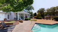 Located at 1011 W Butler Rd in Mauldin, South Carolina, this single-story community clubhouse offers a well-appointed gathering space designed for comfort and recreation. The exterior immediately impresses with a sparkling resort-style swimming pool framed by a generous sundeck, mature shade trees, and meticulously maintained landscaping. Outdoor seating areas provide spots to relax, while a gated walkway lined with towering trees creates a welcoming, park-like approach to the building.

Inside, the main lounge is a standout space featuring a tray ceiling, ceiling fan, and a classic fireplace flanked by wall-mounted artwork and a flat-screen television. Soft, neutral carpeting and expansive grid-pane windows flood the room with natural light, offering views of the lush surroundings. The muted blue-green wall tones lend the interior a calm, sophisticated ambiance throughout.

An adjacent café-style area provides bar-height communal seating along a wooden counter, complemented by a coffee station and additional workspace, making it ideal for casual gatherings or remote work. A bright, carpeted flex room with large windows and greenery views offers additional versatile space. The combination of thoughtful design, abundant natural light, and inviting outdoor amenities makes this clubhouse a true community centerpiece.